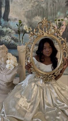 a woman in a white dress holding a gold frame with flowers on the table behind her