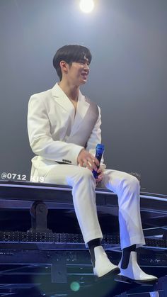 a male in a white suit sitting on a stage and holding a blue bottle with his right hand