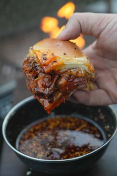 a hand holding up a half eaten sandwich over a bowl of chili and hot sauce