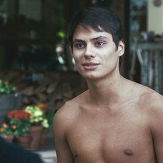 a man with no shirt on standing in front of a potted plant and looking at the camera