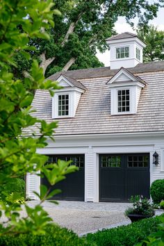 a white house with black garage doors and windows on the top floor is surrounded by greenery