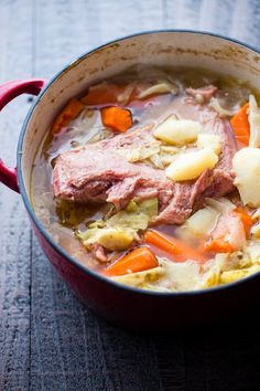 a red pot filled with meat and vegetables on top of a wooden table next to a spoon