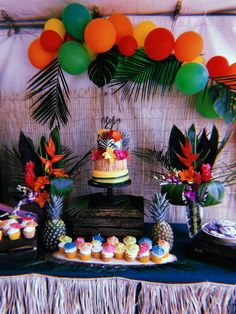 a table topped with lots of cakes and cupcakes next to palm leaf decorations