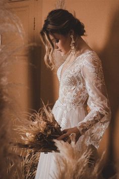 a woman in a white dress standing next to some dry grass and looking down at her bouquet