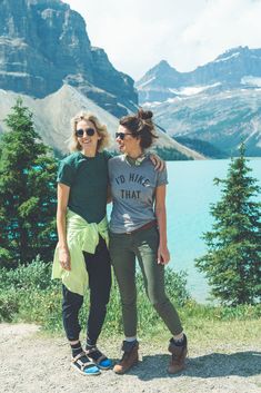 two women standing next to each other on top of a hill near water and mountains