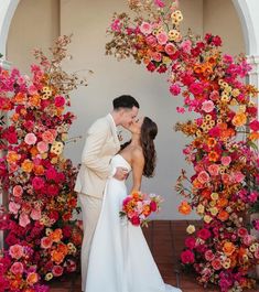 a bride and groom kissing in front of an archway with flowers on the wall behind them