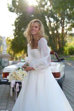 a woman in a white wedding dress standing next to a car and holding a bouquet