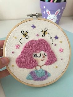 a person holding up a small embroidery hoop with a pink hair girl in the center