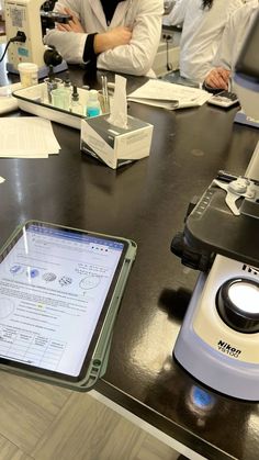 some people are sitting at a table with microscopes and papers on it while one person is holding a tablet