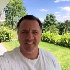 a man in a white t - shirt smiles at the camera while standing on a porch