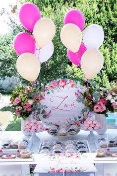 a table topped with lots of cupcakes next to pink and white balloons in the air