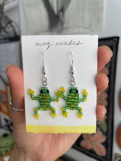 a pair of green and yellow beaded frog earrings on a white card with a black frame