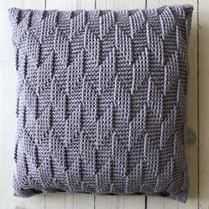 a gray knitted pillow sitting on top of a white wooden floor next to a wall