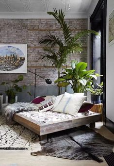 a bed with lots of pillows and plants in the corner next to a brick wall