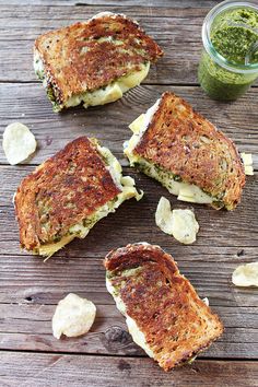 four grilled sandwiches with pesto and cheese on a wooden table next to a jar of pesto sauce