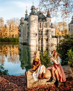 two people sitting on a log in front of a castle with water and trees around them