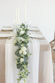 the table is set with white flowers and greenery on it, along with candles