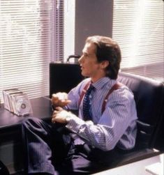 a man sitting in an office chair wearing suspenders and holding his hand to his chest