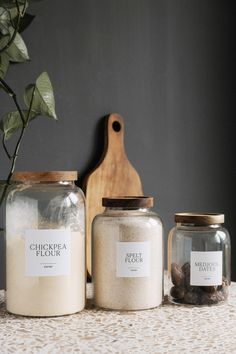 three jars with labels on them sitting on a table next to a cutting board and potted plant