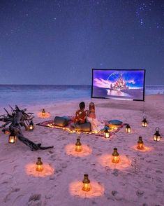 two people are sitting in the sand with lanterns around them, watching a movie at night