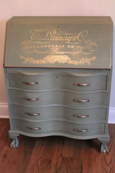 an old dresser painted in gray with gold lettering on the top and bottom, sitting on a hard wood floor