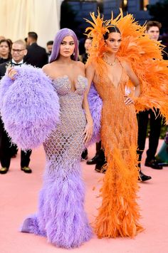 two women dressed in purple and orange feather gowns standing next to each other on a pink carpet