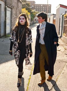 two people walking down the street with makeup painted on their face and hands, one wearing a long black coat