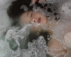 a woman laying down in a bubbley bath with her eyes closed and tongue out