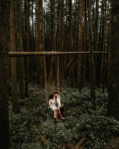 a man and woman are sitting on a swing in the middle of a forest, surrounded by tall trees