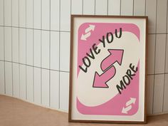 a pink and white sign that says love you more next to a tiled wall with arrows