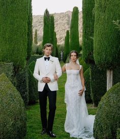 a man and woman dressed in formal wear walking through an archway lined with topiary bushes