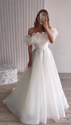 a woman in a white dress taking a selfie with her cell phone while wearing an off the shoulder wedding dress