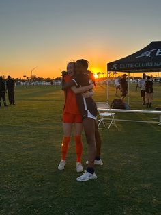 two girls hugging each other on a soccer field at sunset with people in the background