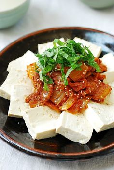 a black plate topped with tofu covered in sauce and garnished with green leaves