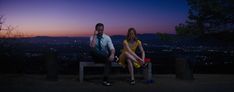 a man and woman sitting on a bench in the evening with city lights behind them