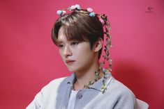 a young man with flowers in his hair wearing a grey sweater and white shirt against a pink background