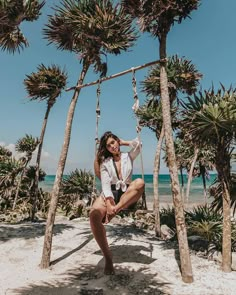 a woman sitting on a swing in front of palm trees at the beach, smiling