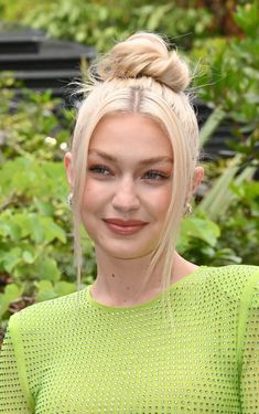 a woman with blonde hair wearing a green top and smiling at the camera while standing in front of bushes