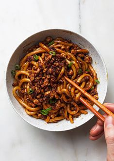 a person holding chopsticks over a bowl of noodles with meat and vegetables in it