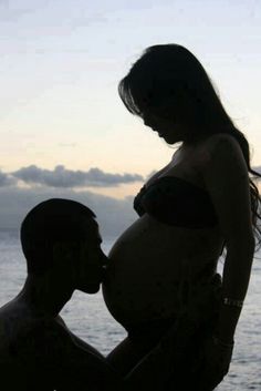 a pregnant woman standing next to a man in front of the ocean with her belly up