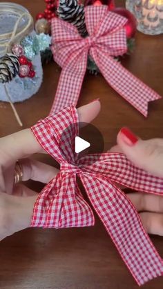 two hands holding red and white bows on top of a wooden table next to pine cones