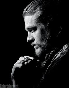 a man with long hair and beard standing in front of a black background holding his hand to his chin