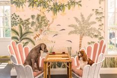 two dogs sitting at a table in front of a wall with palm trees on it