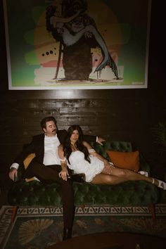 a man and woman are sitting on a green couch in front of a large painting