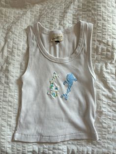 a white tank top with a dolphin embroidered on the front and bottom, sitting on a bed