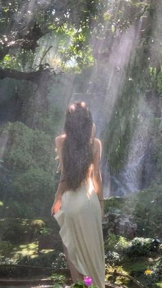 a woman standing in front of a waterfall with her back to the camera and sunlight streaming down