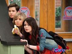 two young people leaning against a counter with a banana in their hand and another person looking at the camera