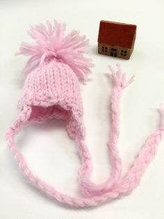 a pink knitted hat next to a small wooden block on a white surface with a tiny house in the background