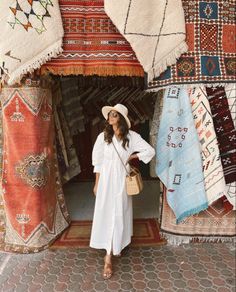 a woman wearing a white dress and hat standing in front of rugs