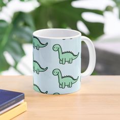 a coffee mug with green dinosaurs on it sitting on a table next to a book